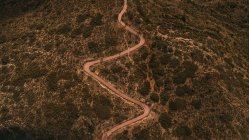 Вид с беспилотника на узкую извилистую сельскую дорогу, проходящую через горную местность с зелеными деревьями и кустарниками в природе в сельской местности — стоковое фото