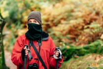 Joven excursionista en ropa de abrigo con cámara fotográfica en el cuello practicando la caminata nórdica en el bosque de otoño y mirando hacia otro lado de ensueño - foto de stock