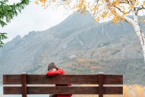 Side view woman in outerwear resting on bench in picturesque autumn park against severe mountain range and calm lake — Stock Photo