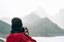 Back view anonymous traveler in outerwear standing on massive rock and taking photo while admiring misty mountain ridge surrounding calm lake on autumn day — Stock Photo