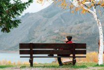 Frau in Oberbekleidung ruht sich auf Bank im malerischen Herbstpark vor schwerer Bergkette und ruhigem See aus und telefoniert — Stockfoto