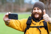 Счастливый взрослый бородатый мужчина-путешественник с большим пальцем вверх делает автопортрет на мобильном телефоне при дневном свете — стоковое фото