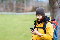Дорослий бородатий чоловік мандрівник переглядає мобільний телефон у денне світло на природі — стокове фото