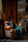 Corpo inteiro de mulher elegante descalça na poltrona olhando para a câmera enquanto descansa no quarto com espelho e interior vintage — Fotografia de Stock