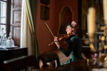 Side view of cheerful female musician touching hair while sitting on armchair in vintage styled room during violin rehearsal — Stock Photo
