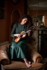 Жінка-музикантка тримає скрипку, сидячи на кріслі у старовинній кімнаті під час репетиції — стокове фото