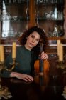 Músico feminino sério com violino olhando para a câmera enquanto se senta à mesa com velas na sala vintage perto de armário de madeira com pratos — Fotografia de Stock