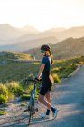 Full body of pensive female in sportswear with helmet standing with bicycle on empty roadway in majestic mountains — Stock Photo