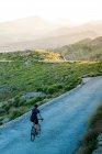 Back view of anonymous female cyclist in casual outfit riding bicycle alone in green highlands with majestic mountains in mist — Stock Photo