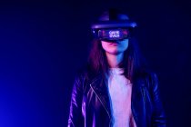 Unrecognizable female wearing modern headset with Game Over inscription while exploring virtual reality in dark room with neon light near wall — Stock Photo