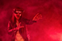 Femme anonyme terrifiée aux bras tendus, coiffée d'un casque moderne hurlant tout en explorant le cyberespace dans une pièce éclairée de néons rouges et de fumée — Photo de stock
