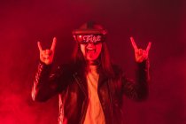 Expressive anonymous female wearing VR goggles with Fire word making horn sign while standing in red neon light with smoke — Stock Photo