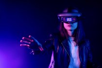 Mulher irreconhecível com braço estendido usando fone de ouvido VR enquanto explora a realidade virtual sob luz de néon azul perto da parede com iluminação do projetor — Fotografia de Stock