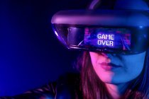Femme méconnaissable portant un casque moderne avec inscription Game Over tout en explorant la réalité virtuelle dans une pièce sombre avec lumière au néon près du mur — Photo de stock