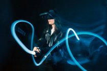 Вид збоку на невизначену жінку, яка носить сучасну гарнітуру VR і грає з контролером руху в синіх неонових вогнях під час вивчення кіберпростору — стокове фото