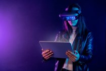 Концентрована анонімна жінка в сучасному гарнітурі VR, що стоїть біля стіни в темній кімнаті з сучасним планшетом в руках під неоновим освітленням — стокове фото