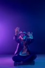 Неузнаваемая женщина с вытянутой рукой в VR гарнитуре во время исследования виртуальной реальности под синим неоновым светом и сидя на полу — стоковое фото