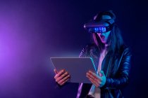 Concentrated anonymous female wearing modern VR headset standing near wall in dark room with contemporary tablet in hands under neon illumination — Stock Photo