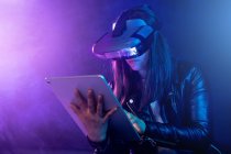 Concentrated anonymous female wearing modern VR headset standing near wall in dark room with contemporary tablet in hands under neon illumination — Stock Photo