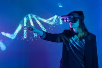 Side view of unrecognizable female with outstretched arm wearing VR headset while exploring virtual reality under blue neon light near wall with projector illumination — Stock Photo