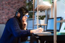 Side view of concentrated female composer in headphones taking notes in notebook while sitting at table with laptop during remote work from home — Stock Photo