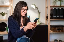 Positive female freelancer in eyeglasses surfing cellphone and sitting near table and shelves with decorations while working remotely from home — Stock Photo