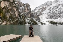 Full body of loving gentle couple hugging each other on wooden pier against Lago di Braies lake surrounded by snowy mountains — Stock Photo