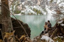 Ganzkörper liebevolle sanfte Paar umarmen einander, während sie auf Wäldern am Pragser See sitzen, umgeben von schneebedeckten Bergen — Stockfoto