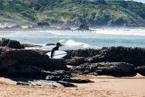 Vista laterale del turista maschio con tavola da surf passeggiando lungo massi sulla costa sabbiosa del mare ondulato schiumoso — Foto stock