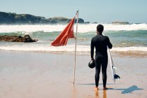 Vista posteriore piena lunghezza dell'uomo anonimo che indossa tuta da surf in piedi vicino alla bandiera sventolante sulla costa lavata da onde schiumose — Foto stock