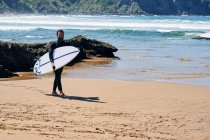 Uomo adulto in muta con tavola da surf sorridente felicemente in piedi sulla riva del mare contro le colline — Foto stock