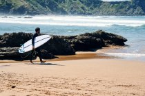 Дорослий чоловік у гідрокостюмі з дошкою для серфінгу, посміхаючись, щасливо стоїть на березі моря проти пагорбів — стокове фото