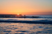 Силуэты людей, плавающих и занимающихся серфингом в волнах моря под ярким солнцем, сияющими на закатном небе — стоковое фото