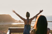 Back view of unrecognizable female taking picture of friend standing with hands raised and enjoying view of sea at sunset — Stock Photo