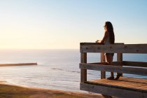 Vista lateral de cuerpo completo de turista femenina parada en terraza de madera y admirando pintoresco paisaje de mar sin fin al atardecer - foto de stock