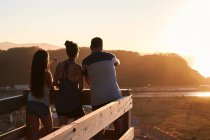 Visão traseira de corpo inteiro de turistas em pé na plataforma de madeira e admirando o mar calmo durante o pôr do sol — Fotografia de Stock