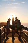 Touristes souriants appuyés sur une rampe en bois et prenant autoportrait contre le coucher du soleil ciel sur la mer calme — Photo de stock