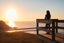 Visão lateral de corpo inteiro de turista feminino em pé no terraço de madeira e admirando a paisagem pitoresca do mar sem fim ao pôr do sol — Fotografia de Stock