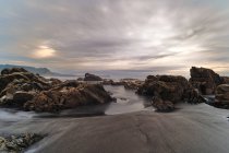 Vue pittoresque des rochers couverts de mousse sur la plage de sable fin de la mer sous le ciel nuageux du coucher du soleil — Photo de stock