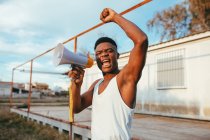 Young angry African American male in undershirt with speaker yelling with raised arm while looking at camera — Stock Photo