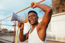 Jovem afro-americano irritado macho em camiseta com alto-falante gritando com braço levantado enquanto olha para a câmera — Fotografia de Stock