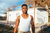 Young masculine African American male in undershirt standing near platform and looking at camera against house — Stock Photo