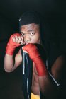 Young strong African American sportsman in boxing hand wraps working out and looking at camera in building — Stock Photo