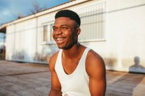 Happy African American male in undershirt with modern haircut looking forward against building in sunlight — Stock Photo