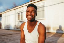 Happy African American male in undershirt with modern haircut looking at camera against building in sunlight — Stock Photo