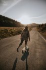 Вид ззаду безлика спортивна жінка в модному одязі верхи на крейсерській дошці вздовж порожньої асфальтової дороги в літній сільській місцевості в сонячний день — стокове фото