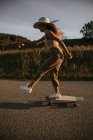 Seitenansicht Sportliche Frau in trendiger Kleidung fährt Cruiser Board entlang leerer Asphaltstraße in sommerlicher Landschaft an sonnigem Tag — Stockfoto