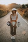 Full body content young female in casual wear and summer hat holding cruiser skateboard and looking at camera while standing on empty asphalt road in rural area at sunset — Stock Photo