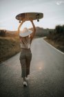 Junge Frau in Freizeitkleidung und Sommermütze hält Cruiser-Skateboard in der Hand und schaut weg, während sie bei Sonnenuntergang auf einer leeren Asphaltstraße im ländlichen Raum steht — Stockfoto