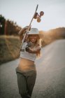 Вміст молодої жінки в повсякденному вбранні і літньому капелюсі тримає крейсер скейтборд і дивиться на камеру, стоячи на порожній асфальтовій дорозі в сільській місцевості на заході сонця — стокове фото